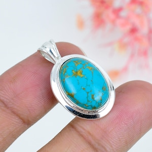 Puede incluir: Un colgante ovalado con una piedra turquesa brillante con vetas de matriz de color marrón dorado, engastado en un marco de plata pulida con una anilla de plata a juego en la parte superior, con una superficie cabujón lisa y brillo.