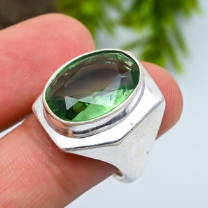 May include: A silver ring with a large, oval-cut green gemstone. The gemstone displays a gradient of green shades. The ring has a geometric, angular design around the stone, with a polished finish.