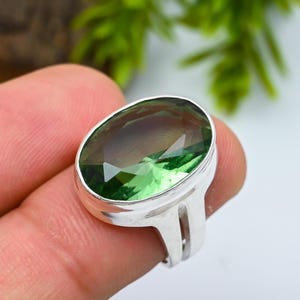 May include: A silver ring featuring a large, oval-cut, faceted gemstone that transitions in color from a deep olive green at one end to a lighter, translucent lime green at the other, set in a polished, wide, split-shank metal band.
