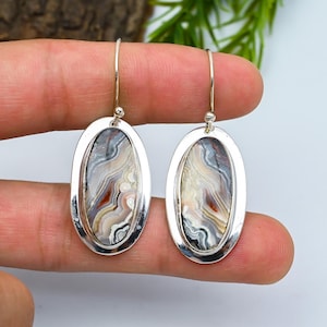 May include: A pair of silver oval drop earrings. Each earring showcases a polished stone with a swirling pattern of grey, white, beige, and red tones. The earrings are suspended from a simple silver hook.