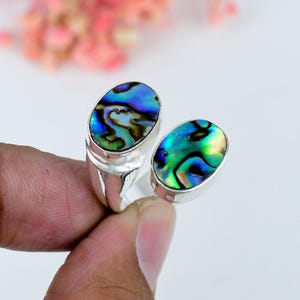 May include: A silver ring featuring two oval-shaped abalone shell inlays. The shell displays iridescent swirls of blue, green, and purple colors. The metal band is polished and smooth, holding the two distinct shell settings in a bypass design.