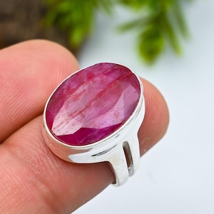 Puede incluir: Un anillo de plata con una gran gema de rubí de corte ovalado. El rubí muestra un tono rosa-rojo vibrante con patrones internos visibles. La banda del anillo es un diseño simple de plata pulida.