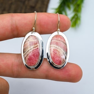 Pode incluir: Um par de brincos ovais com moldura prateada. Cada brinco apresenta uma pedra rosa e branca listrada. Os brincos pendem de ganchos prateados. Uma peça de joalharia.