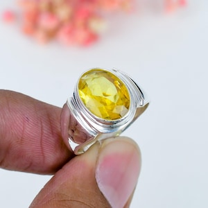 Peut inclure: Une bague en argent ornée d'une grande pierre jaune facettée de taille ovale, sertie dans une monture en métal poli. L'anneau présente une structure lisse et solide qui s'affine légèrement vers l'arrière, avec une finition brillante et réfléchissante.