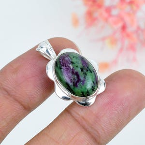 Pode incluir: Um pingente de pedra preciosa rubi zoisita de formato oval engastado em uma moldura de prata polida. A pedra apresenta um padrão manchado de magenta profundo e verde floresta com inclusões pretas. O engaste de prata tem uma borda festonada.