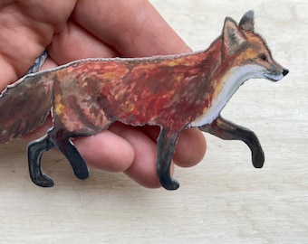 May include: A hand-painted paper cutout of a red fox with a white tip on its tail. The fox is walking with its head turned to the right. The cutout is made of paper and has a textured surface.