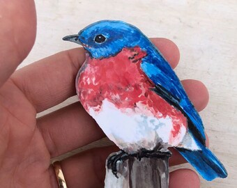 May include: A hand-painted wooden bird figurine. The bird has blue wings and head, a red breast, and a white belly. It is perched on a wooden post. The bird is a charming decorative item.