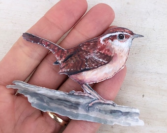 May include: A paper cut-out of a brown and white bird with a long tail perched on a branch. The bird has a white breast and a brown back with a white stripe across its eye.