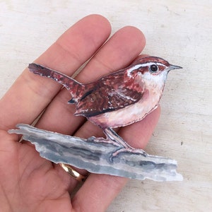 Può includere: Un ritaglio di carta di un uccello marrone e bianco con una lunga coda appollaiato su un ramo. L'uccello ha un petto bianco e una schiena marrone con una striscia bianca sopra l'occhio.