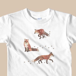 May include: White t-shirt with a graphic of three red foxes walking on a white background with small black paw prints.