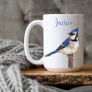 May include: White ceramic mug featuring a watercolor illustration of a blue jay perched on a branch. The name "Jamie" is written in blue script. The mug has a large handle and is set against a neutral background.