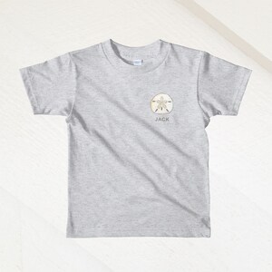 K&ouml;nnte beinhalten: Graues T-Shirt mit kurzen &Auml;rmeln, einem Sanddollar-Motiv und dem Namen "JACK" darunter.