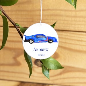 May include: A round white ornament with a blue car painted on it. The ornament has the name 'ANDREW' and the year '2021' written on it.