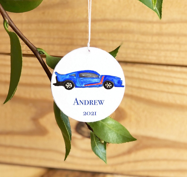 May include: A round white ornament with a blue car painted on it. The ornament has the name 'ANDREW' and the year '2021' written on it.
