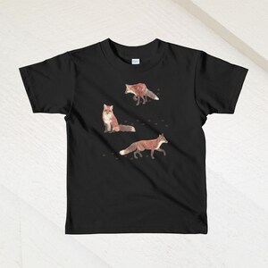 May include: Black t-shirt with a design of three red foxes on a white background. The foxes are in different poses, one is sitting, one is walking, and one is running. There are small brown paw prints scattered around the foxes.