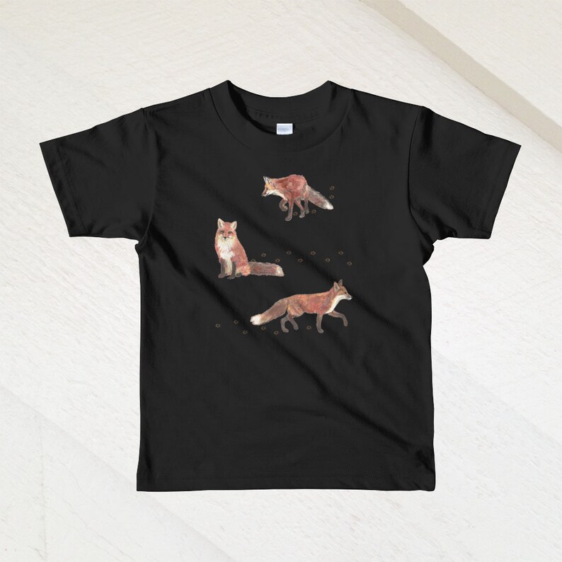 May include: Black t-shirt with a design of three red foxes on a white background. The foxes are in different poses, one is sitting, one is walking, and one is running. There are small brown paw prints scattered around the foxes.