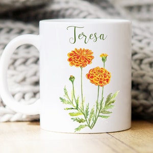 May include: White ceramic mug with a handle, featuring a watercolor illustration of orange marigolds and the name "Teresa" printed in black cursive.