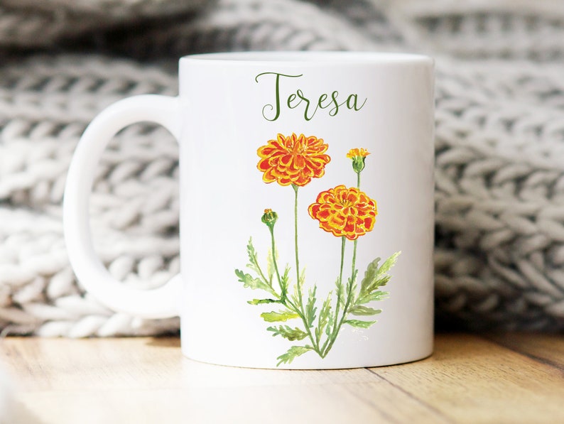 May include: White ceramic mug with a handle, featuring a watercolor illustration of orange marigolds and the name "Teresa" printed in black cursive.