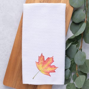 Puede incluir: Toalla de cocina blanca con un diseño de una sola hoja de arce naranja y amarilla.