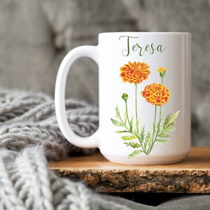 May include: White ceramic mug with a floral design featuring orange and yellow marigolds. The mug has the name "Teresa" written on it.