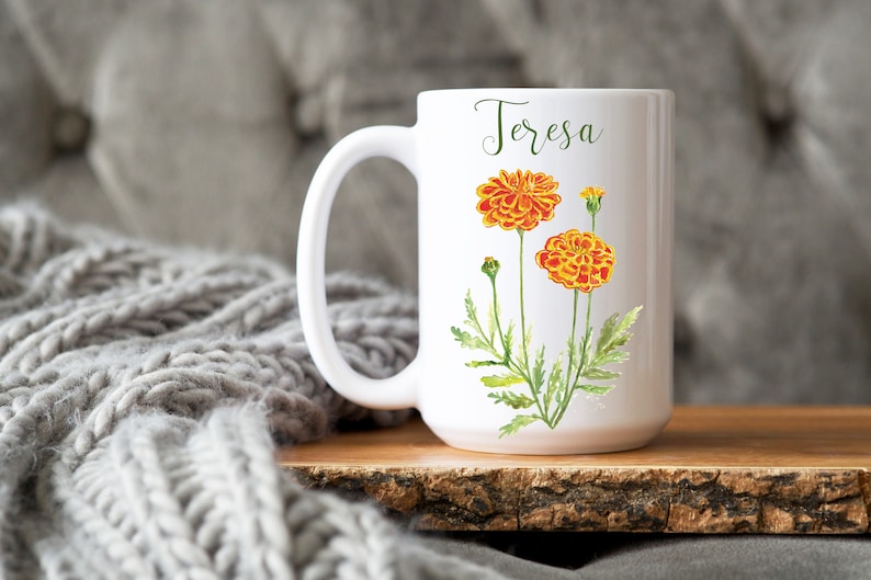 May include: White ceramic mug with a floral design featuring orange and yellow marigolds. The mug has the name "Teresa" written on it.