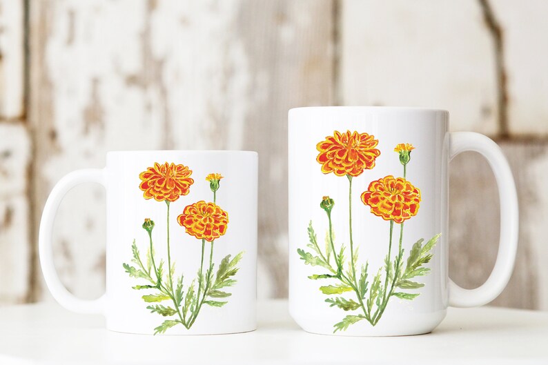 May include: Two white ceramic mugs with a floral design. The mugs feature orange and yellow marigold flowers with green stems and leaves.