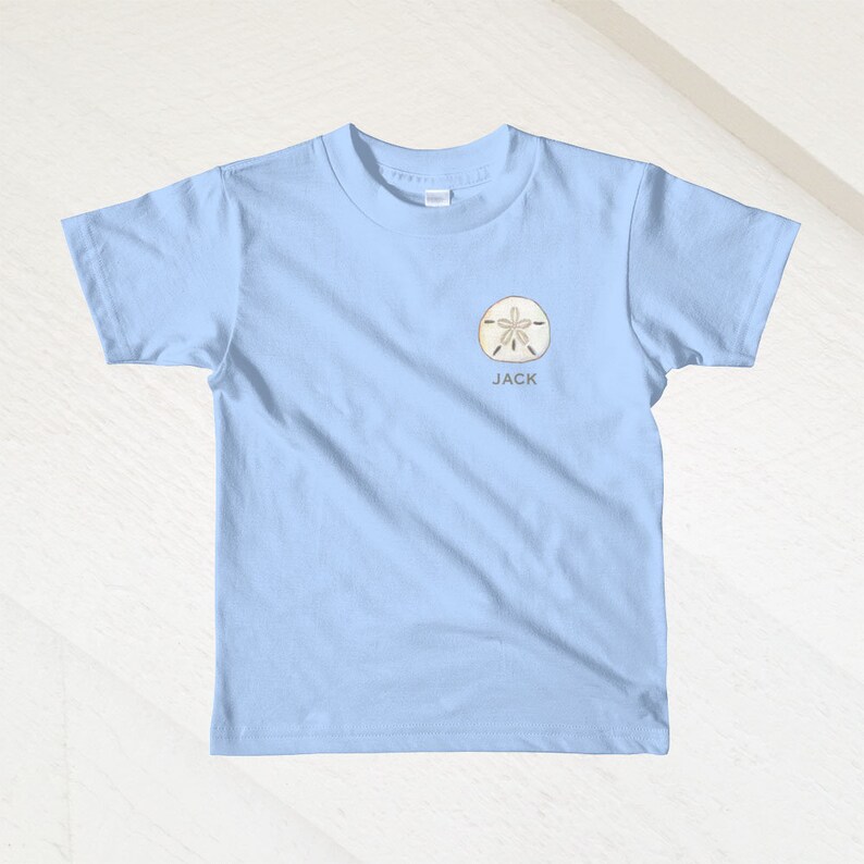 K&ouml;nnte beinhalten: Hellblaues T-Shirt mit einem Sanddollar-Motiv und dem Namen "JACK" auf der Vorderseite.