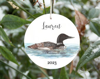 Könnte beinhalten: Ein weißer runder Weihnachtsbaumschmuck mit einer Aquarell-Illustration eines Haubentauchers, der in blauem Wasser schwimmt. Der Schmuck ist mit dem Namen "Lauren" und dem Jahr "2023" personalisiert.