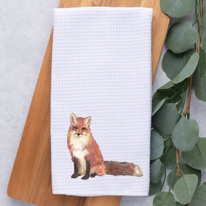 May include: White waffle weave kitchen towel with an embroidered image of a red fox sitting with its tail curled behind it.