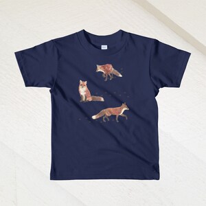 May include: A navy blue children's t-shirt with a graphic of three red foxes on a white background. The foxes are in different poses, one is walking, one is sitting, and one is running. There are small brown paw prints scattered around the foxes.