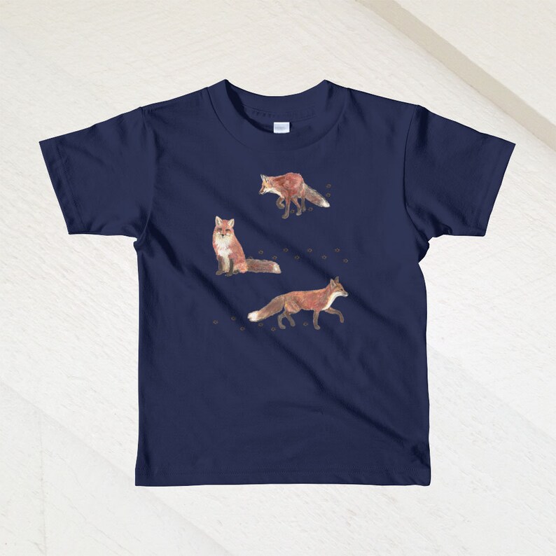 May include: A navy blue children's t-shirt with a graphic of three red foxes on a white background. The foxes are in different poses, one is walking, one is sitting, and one is running. There are small brown paw prints scattered around the foxes.
