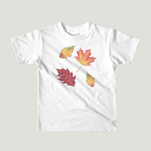以下が含まれることがあります： 白いTシャツに、赤、オレンジ、黄色の4枚の秋の葉のデザインが施されています。葉は木から落ちています。