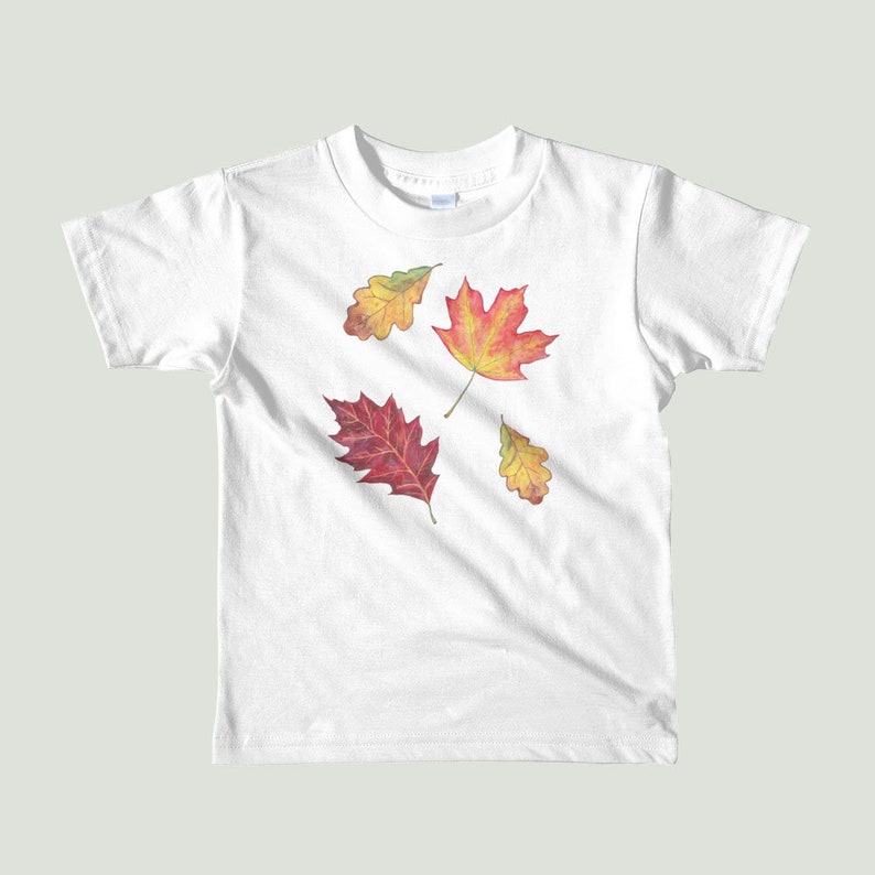 以下が含まれることがあります： 白いTシャツに、赤、オレンジ、黄色の4枚の秋の葉のデザインが施されています。葉は木から落ちています。