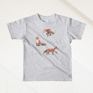 May include: A heather grey t-shirt with a design of three foxes and paw prints. The foxes are brown and orange with white accents.