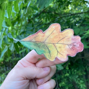 May include: A hand holding up a paper oak leaf painted in shades of green, yellow, and red.