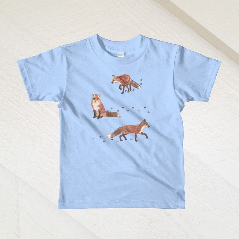 May include: A light blue t-shirt with a graphic of three red foxes walking on a white background. The foxes are walking in different directions and have black eyes and noses.
