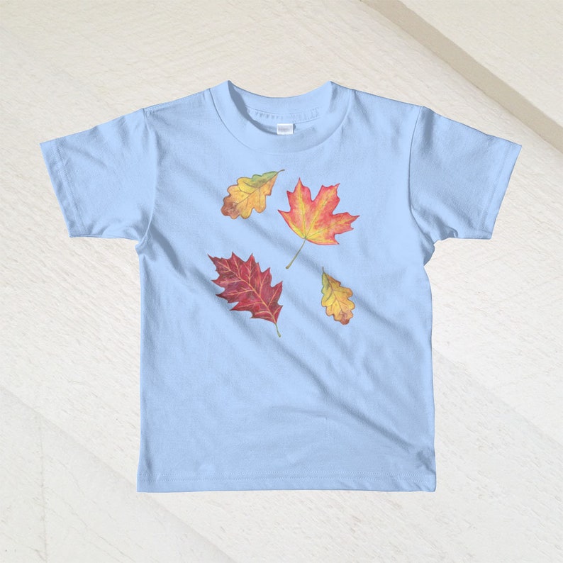 以下が含まれることがあります： 薄い青色のTシャツに、赤、オレンジ、黄色の秋の葉が4枚デザインされています。葉はTシャツの前側に散らばっています。
