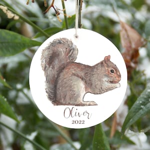 May include: A white round Christmas ornament with a watercolor illustration of a gray squirrel holding a nut. The ornament has the name "Oliver" and the year "2022" printed on it.
