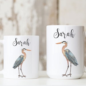 May include: Two white ceramic mugs with a blue heron design and the name "Sarah" printed on each. The mugs are on a white surface.