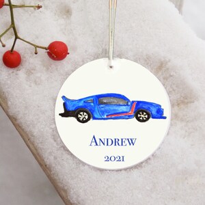 May include: A round white Christmas ornament with a blue car painted on it. The car has a red stripe and black wheels. The ornament also has the name 'ANDREW' and the year '2021' written on it.