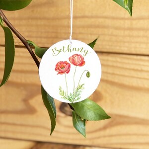 May include: A white round ornament with the name Bethany written in cursive script. The ornament features two red poppy flowers with green stems and leaves.