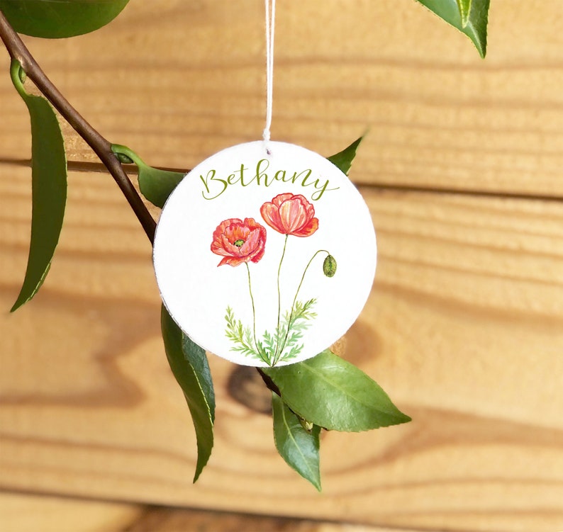 May include: A white round ornament with the name Bethany written in cursive script. The ornament features two red poppy flowers with green stems and leaves.
