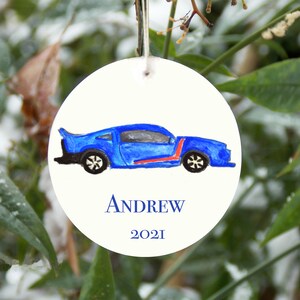 May include: A round white ornament with a blue car drawn on it. The car has a red stripe and black wheels. The ornament has the name 'ANDREW' and the year '2021' written on it.