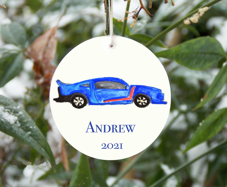 May include: A round white ornament with a blue car drawn on it. The car has a red stripe and black wheels. The ornament has the name 'ANDREW' and the year '2021' written on it.