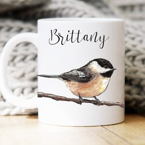 May include: White ceramic mug with a black and white illustration of a chickadee perched on a branch. The mug has the name "Brittany" written in black cursive on the front.