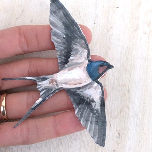 Op de afbeelding: Een aquarel van een zwaluw in de vlucht. De vogel is geschilderd in tinten grijs, wit en blauw, met een zwarte snavel en een blauwe kop.