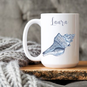 May include: White ceramic mug with a blue seashell design and the name "Laura" written in cursive on the front.