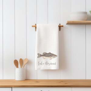 May include: White waffle weave kitchen towel with a brown bass fish illustration and the text "Lake Norman" printed in black.