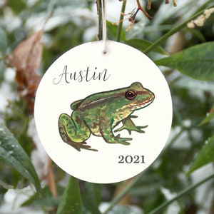 May include: A white round ornament with a green frog illustration and the name "Austin" and the year "2021" printed on it.