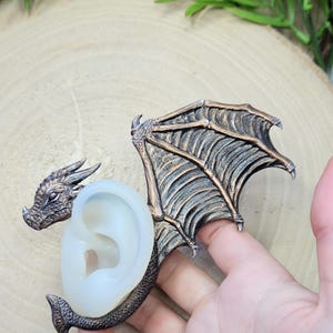 May include: A silicone mold for making a dragon ear cuff. The mold is clear and shows the detailed design of the dragon, including its wings, head, and tail.